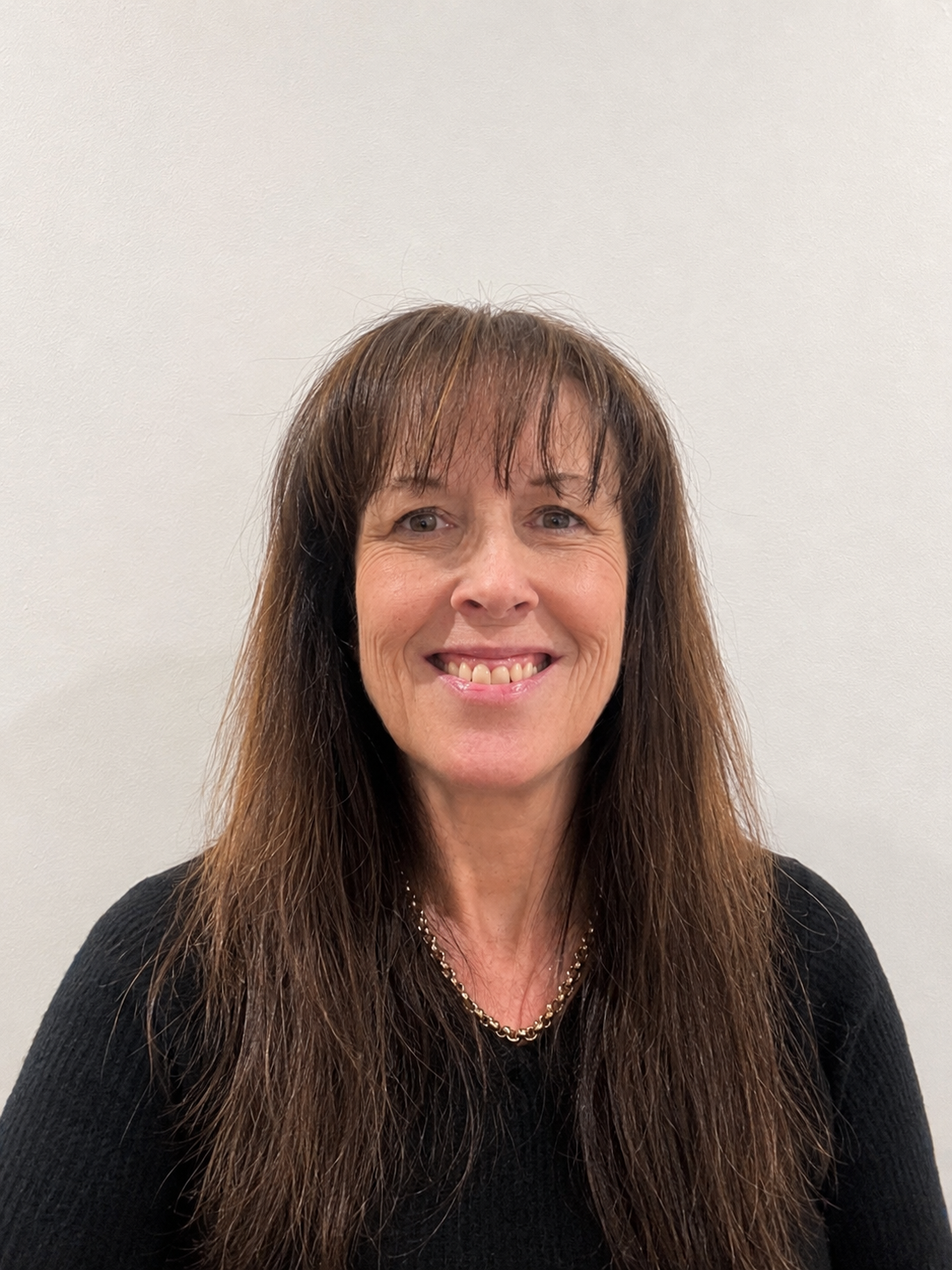 A middle-aged woman with long brown hair, smiling, wearing a black top and a gold chain necklace, in front of a plain white wall.