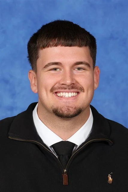 A young man with short brown hair and a goatee, wearing a black jacket over a white shirt and black tie, smiling against a blue background.