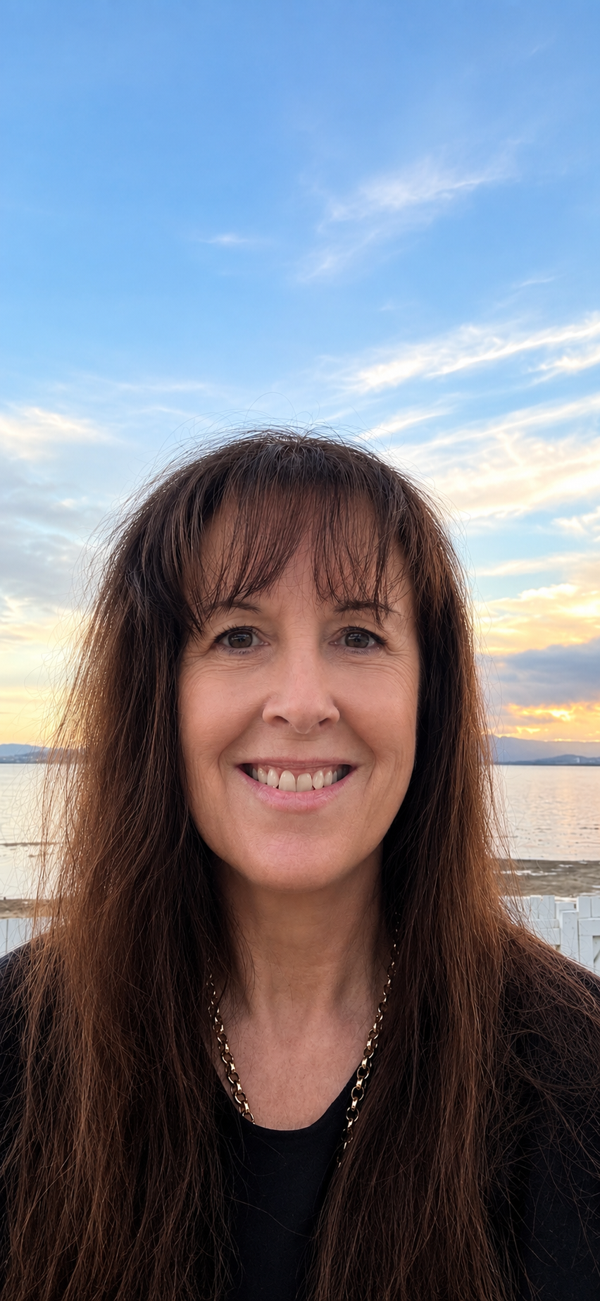 A woman smiling outdoors at sunset with a body of water and a partly cloudy sky in the background.