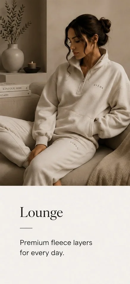 A woman wearing a cream-colored fleece lounge set, sitting on a beige sofa in a cozy living room with minimalist decor, including a white vase with green branches, a lit candle, and books.