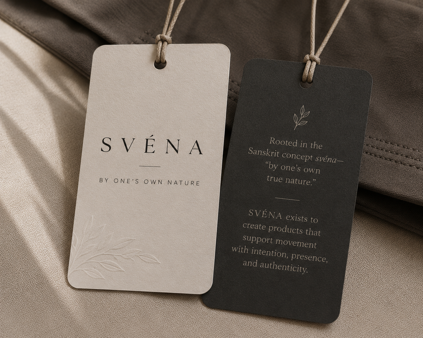 Two product tags hanging from a fabric surface, one in cream and one in dark gray, with text about the brand Svéna and its Sanskrit roots.