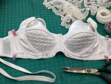 Handcrafted white satin underwire bra in progress on a sewing table, featuring quilted foam cups, lace details, and sewing tools.