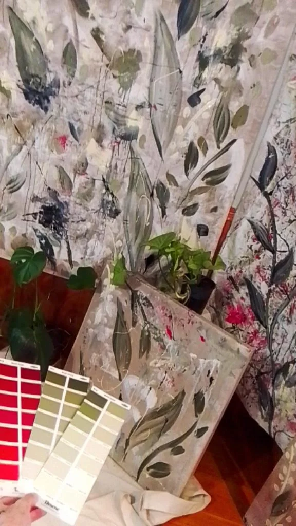 Interior space with floral-patterned wallpaper, various potted houseplants, color swatches, and a framed artwork with a similar floral pattern leaning against the wall.