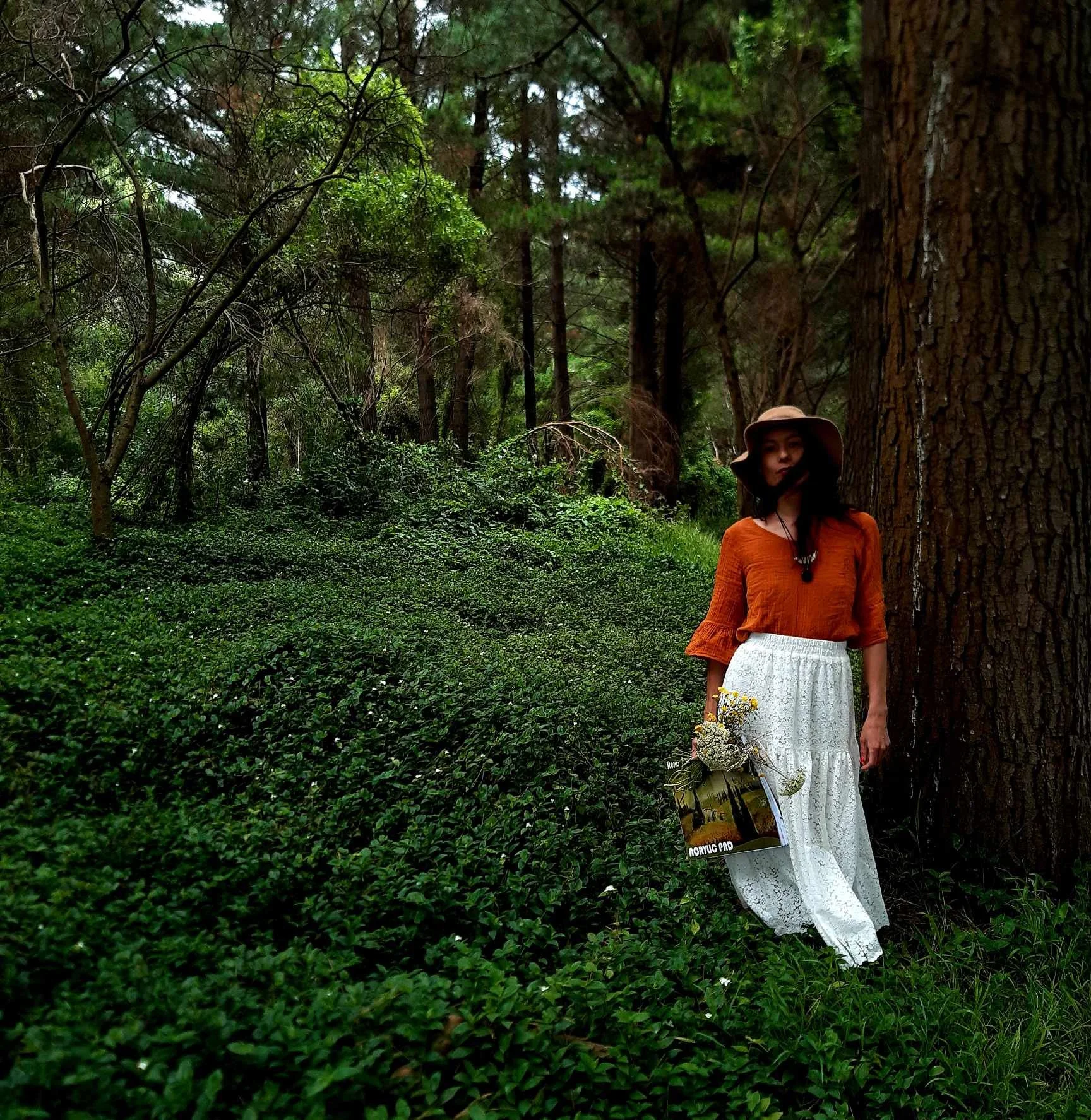 A young woman with dark hair wearing an orange top, white lace skirt, and a wide-brimmed hat, standing in a dense forest with tall trees and green foliage, holding a bouquet of flowers and a bag with a magazine on it.