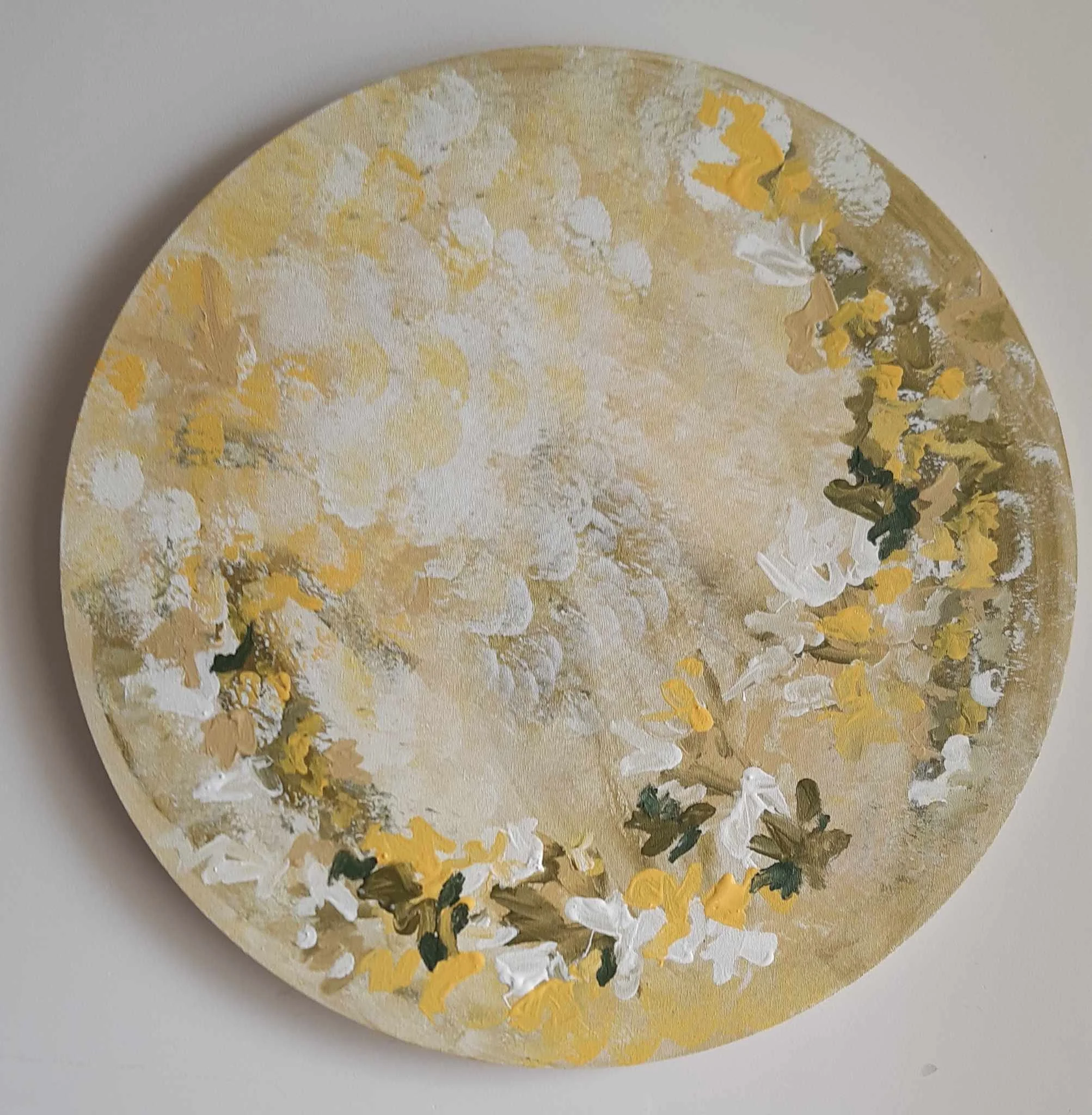 Close-up of a round artwork with a textured yellow and white paint surface and green accents, set against a plain light grey background.