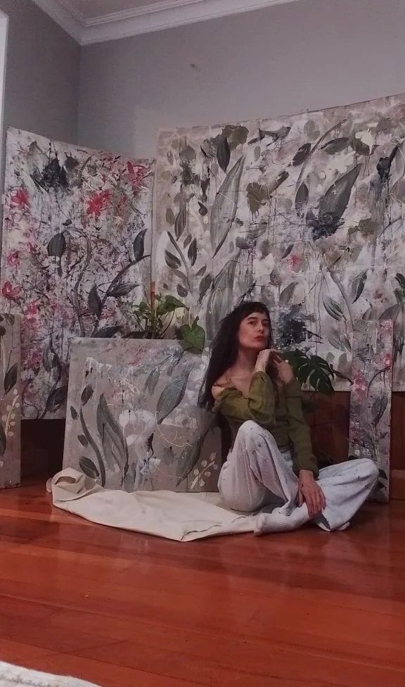 Young woman sitting on a wooden floor among large abstract floral and leaf-patterned canvases in a room with gray walls and crown molding.