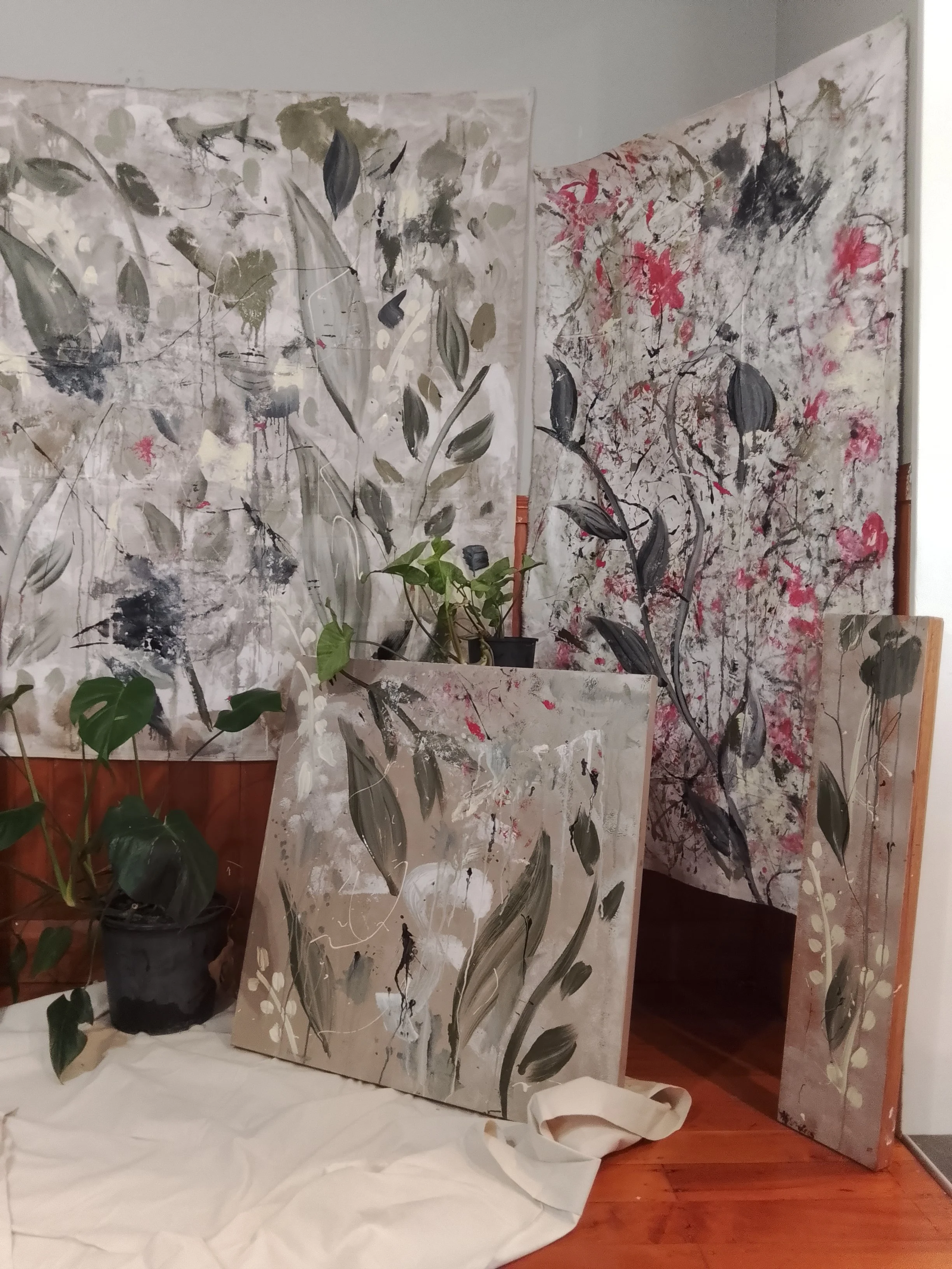 Multiple large abstract paintings with botanical and nature-inspired designs arranged in a room, with some paintings leaning against the wall and others standing upright, along with potted plants and a white cloth on the floor.