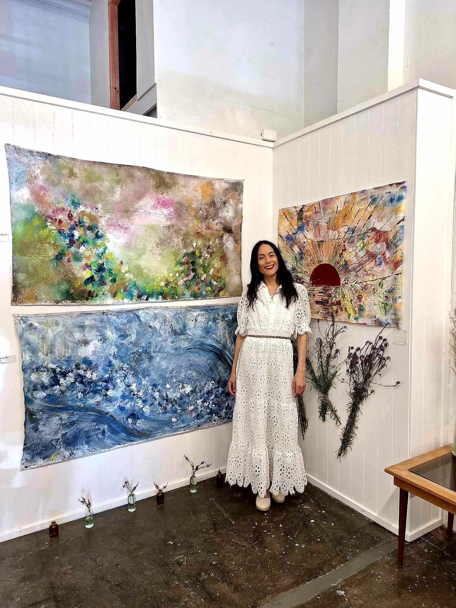 A woman wearing a white lace dress standing in front of two abstract paintings in an art gallery. The paintings feature vibrant colors and textured brushstrokes; one depicts a cityscape and the other an explosion of colors with a red circle. There ar