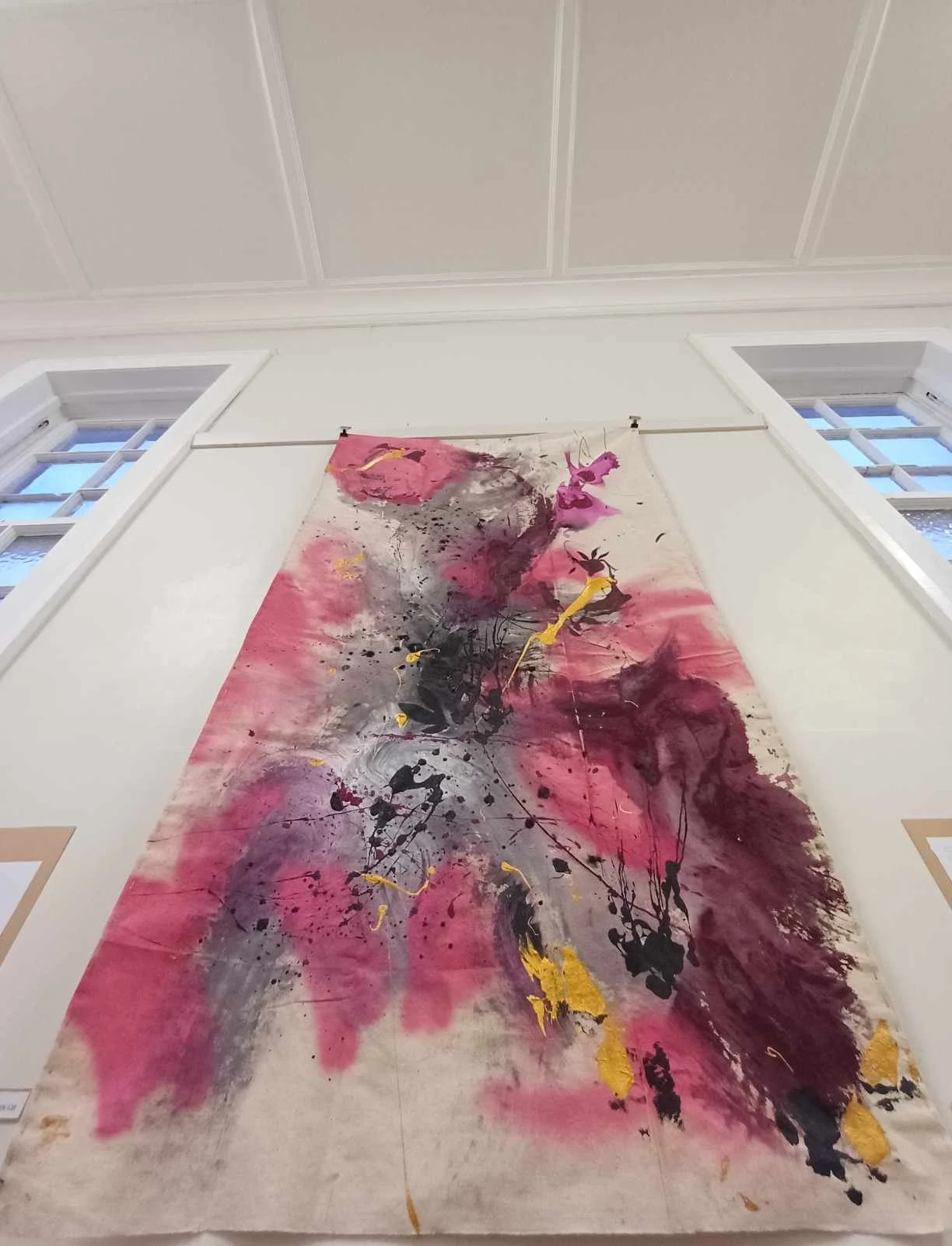 An abstract painting with pink, black, gray, and yellow splashes on a vertical canvas, hanging on a wall near two windows.