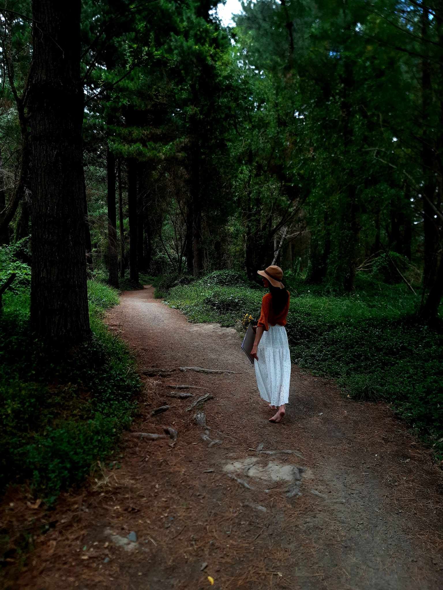 A woman in a white dress and sunhat walking barefoot on a dirt trail in a lush green forest.