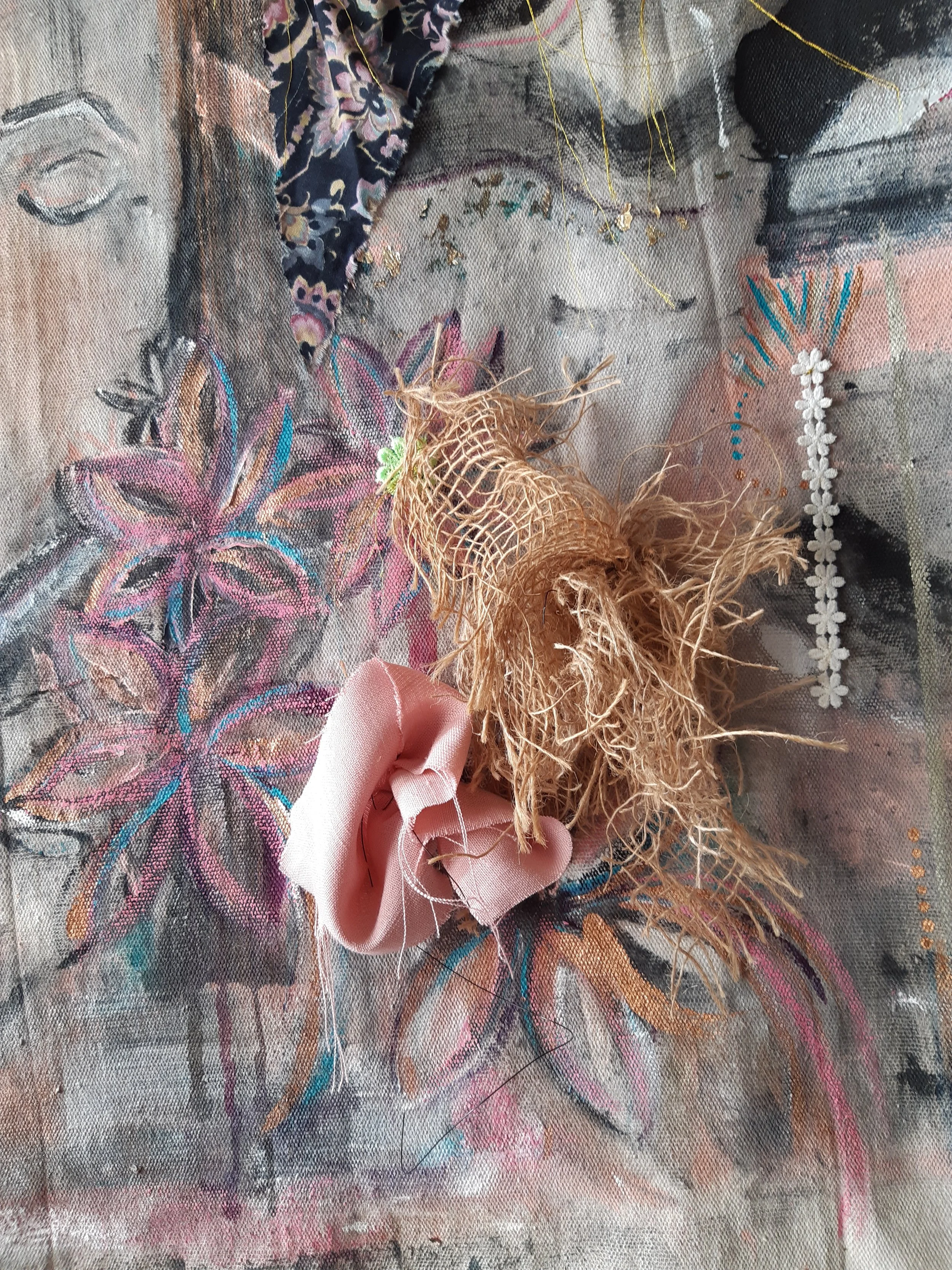 Close-up of textile art with colorful embroidered flowers, gold thread accents, and a handmade fabric flower attached, with a person's finger holding a small pink ribbon.
