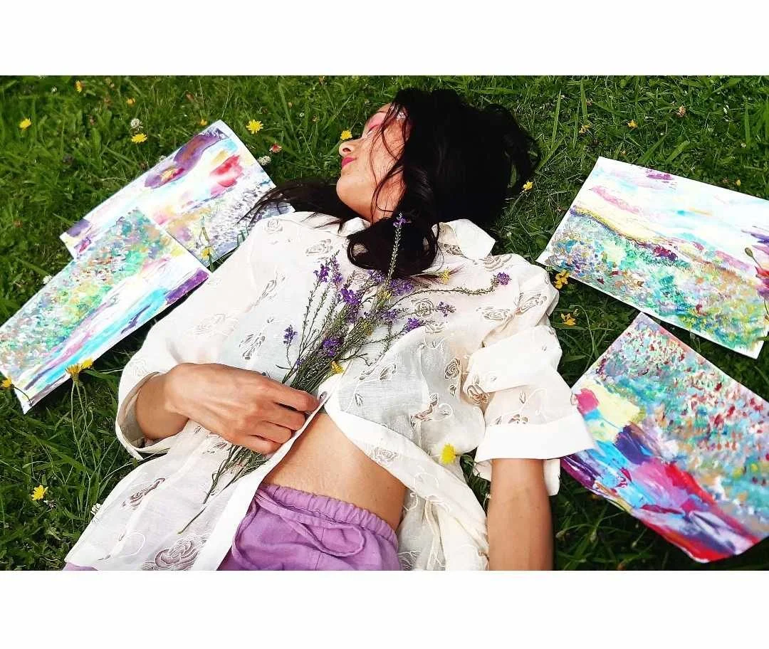 A woman with dark hair lying on green grass surrounded by colorful abstract paintings and holding a bunch of lavender flowers.