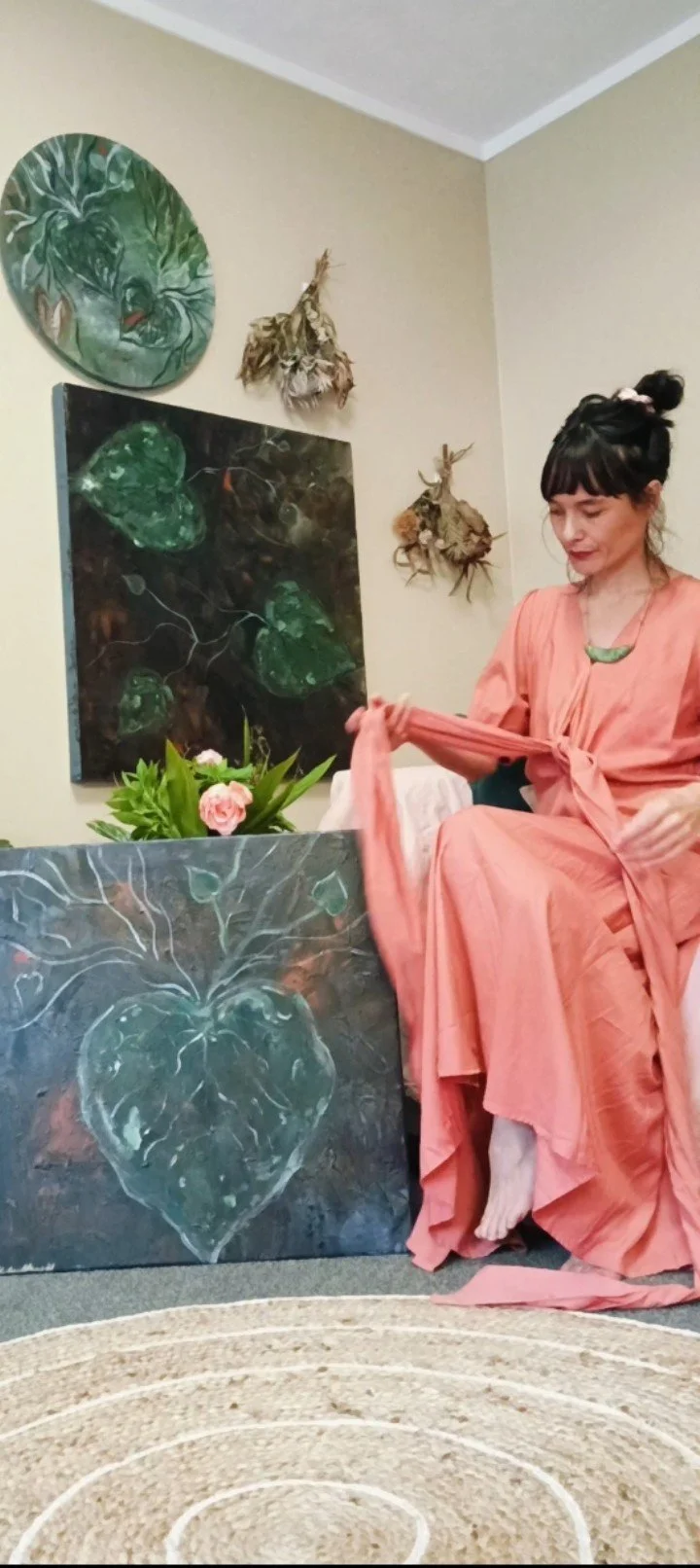 Woman in pink dress sitting next to a painting of a heart with leaf patterns, holding the fabric of her sleeve, in a room with artwork and dried plants on the walls.