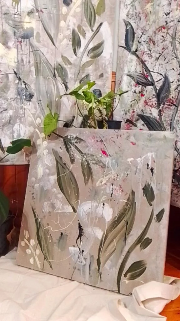 A canvas with abstract floral and leaf patterns leaning against a wall with similar floral decor. There are potted plants nearby, and the canvas features white, black, and green brushstrokes.