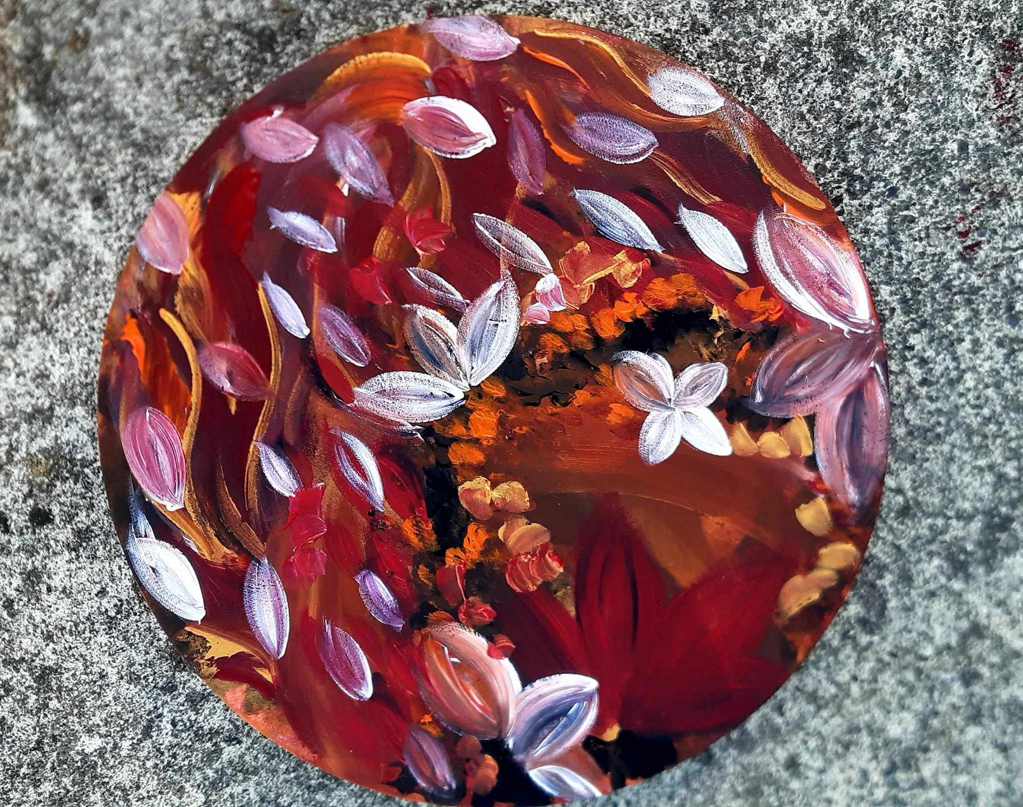 A painted rock with colorful abstract designs including purple, white, orange, and black on a rough gray surface.