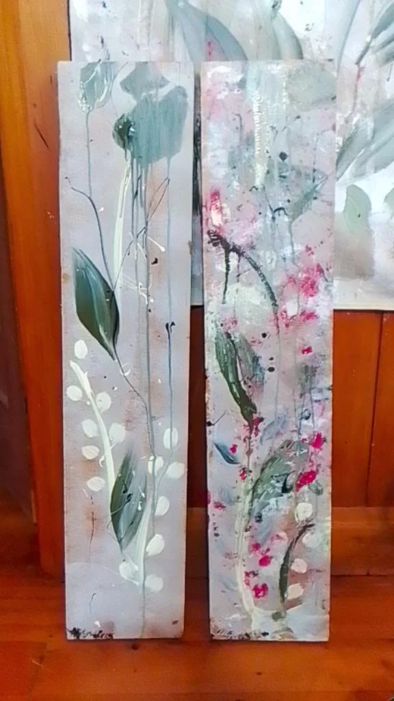 Two abstract floral paintings on tall wooden panels, featuring green leaves, white dots, and splashes of pink, blue, and black paint.