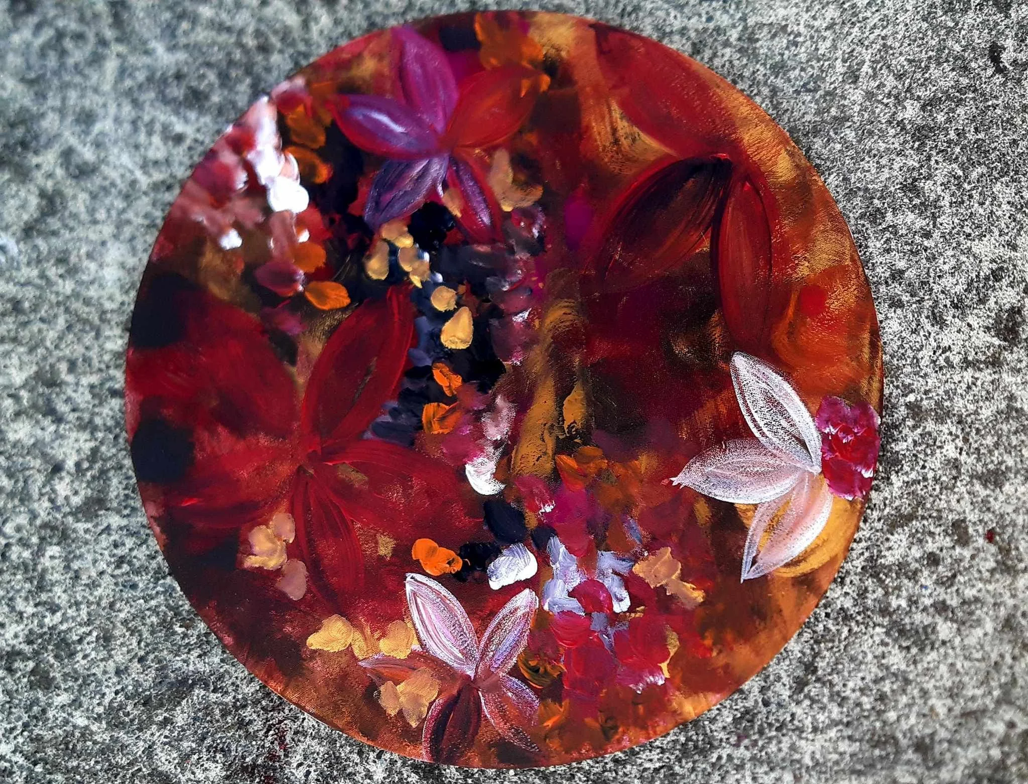 A circular painting with a red background decorated with small, colorful flower petals and white leaf shapes, on a gray speckled surface.
