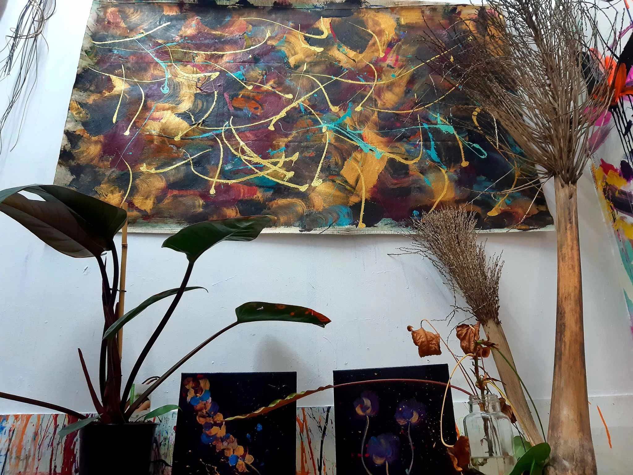 An abstract painting with swirling dark background, splashes of bright colors like gold, teal, pink, and orange, and gold and teal drips and lines across the canvas. The painting is displayed on a wall with surrounding plants and other artworks.