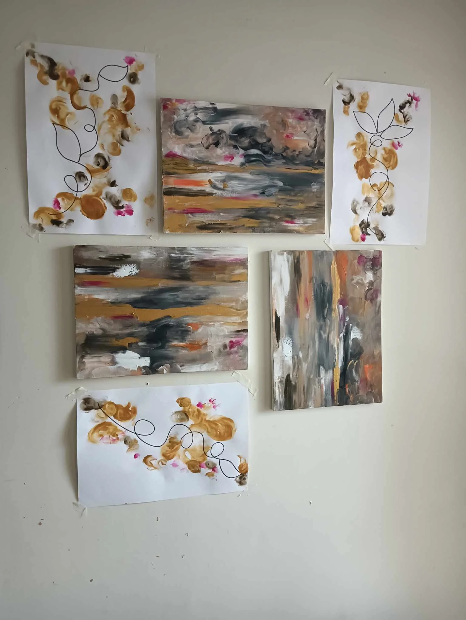 A wall display featuring five abstract landscape paintings with earthy tones and two decorative paper artworks with black line drawings and splotches of brown, pink, and black paint.