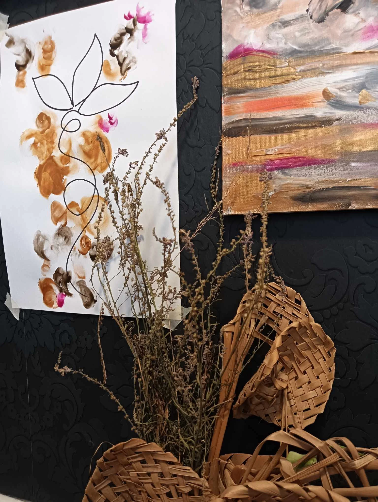 A floral art arrangement featuring dried plants in a woven basket, background paintings with abstract and botanical themes, and a decorative black surface with a raised pattern.