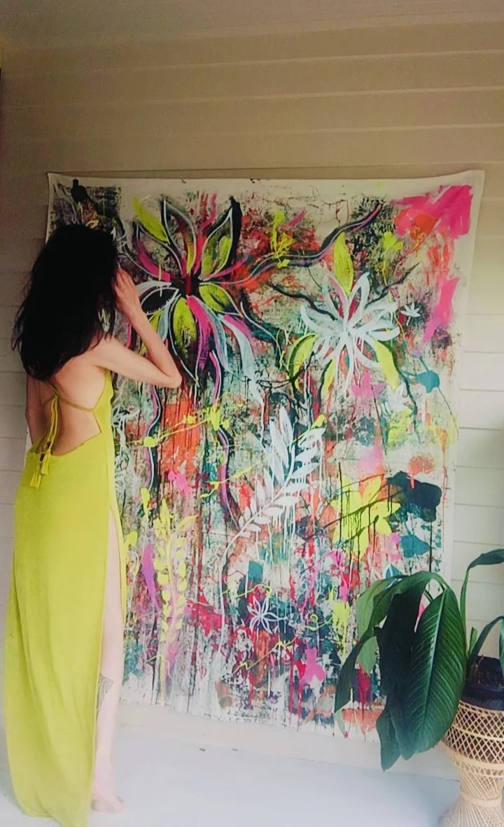 A woman with long dark hair standing next to a colorful abstract floral painting on a wall. She is wearing a bright yellow gown with side slits.