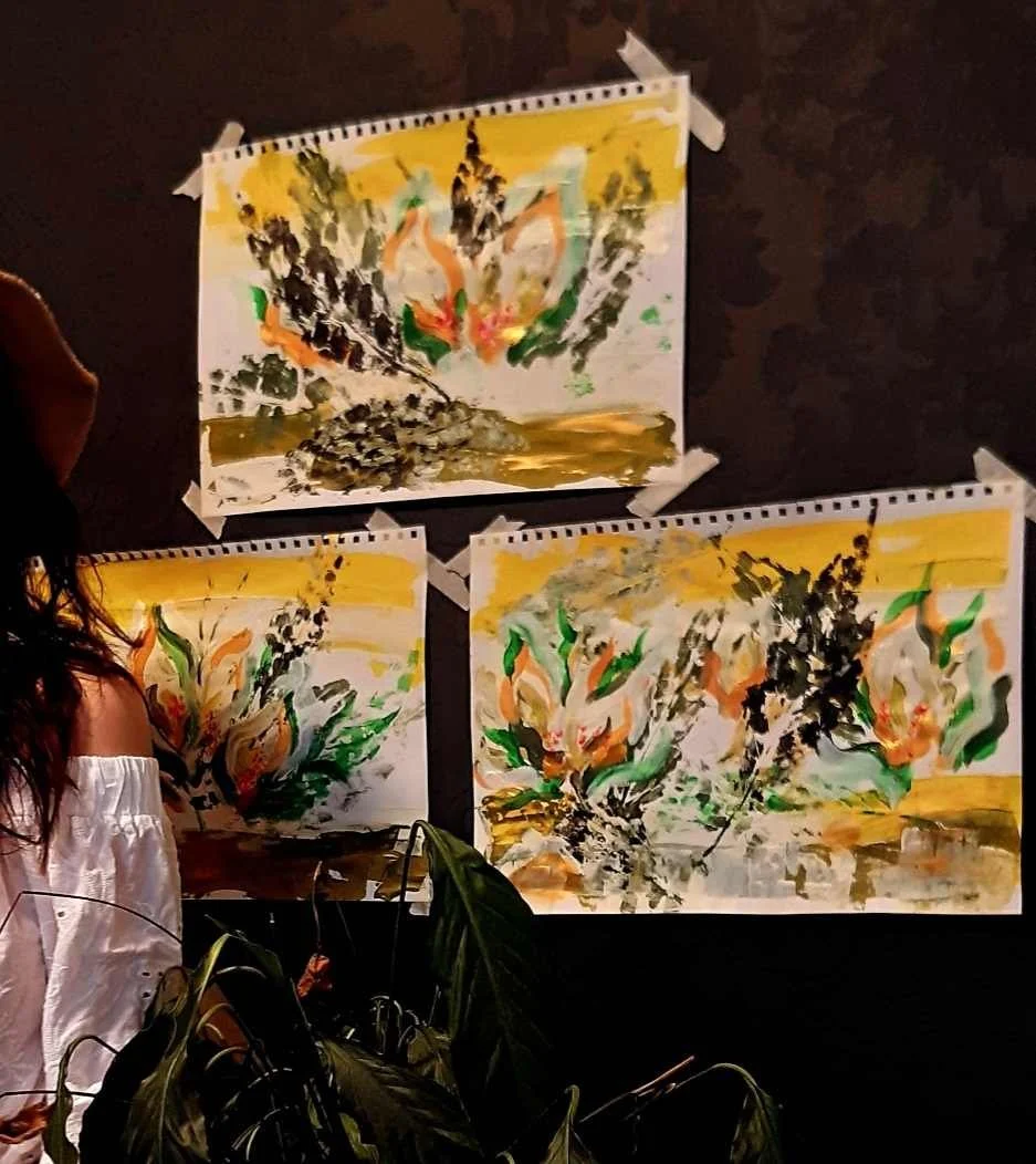 Three abstract paintings featuring bold brush strokes and vibrant colors such as green, orange, and black, displayed on a black wall with white mounting tape, and a woman with long dark hair and an off-the-shoulder white top standing nearby in a diml