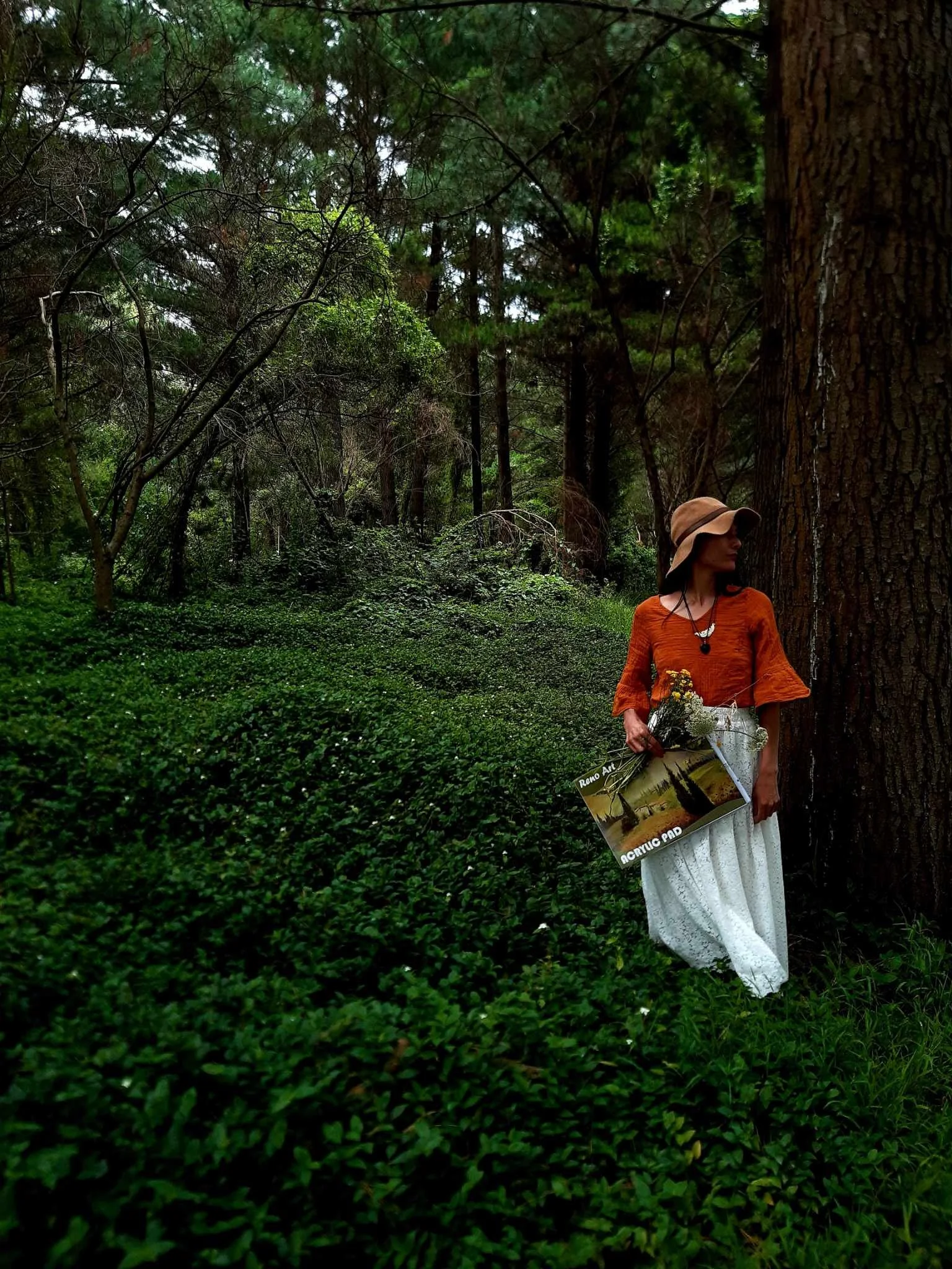 A woman with a wide-brimmed hat and orange top standing in a lush green forest, holding a book or magazine with an image of a park or nature scene on the cover. She is slightly leaning against a large tree trunk.