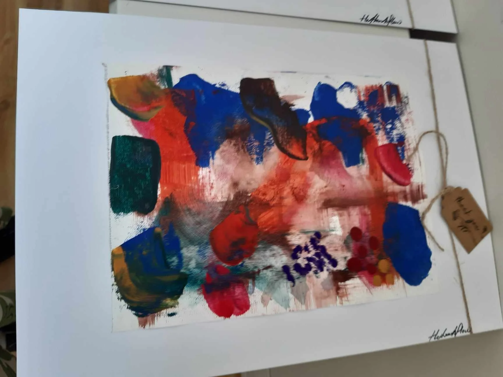 Abstract colorful painting with vivid shades of red, blue, orange, and green, featuring various shapes and textures, framed and laying on a light-colored surface.