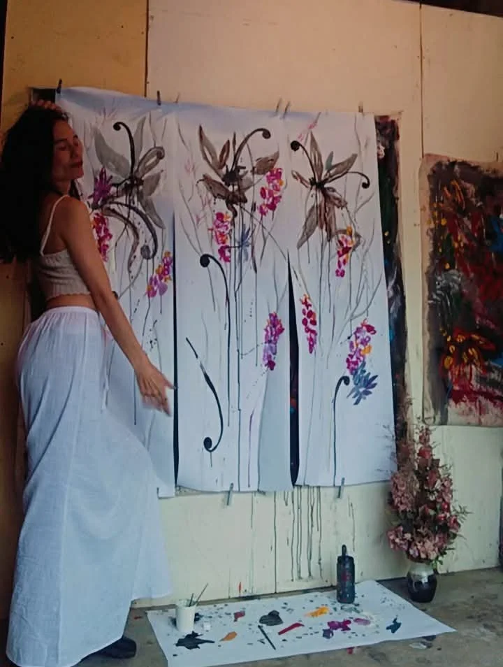 Woman in white skirt and tank top standing next to a large canvas painting of stylized flowers with pink and purple accents, in an art studio with paintings and art supplies.
