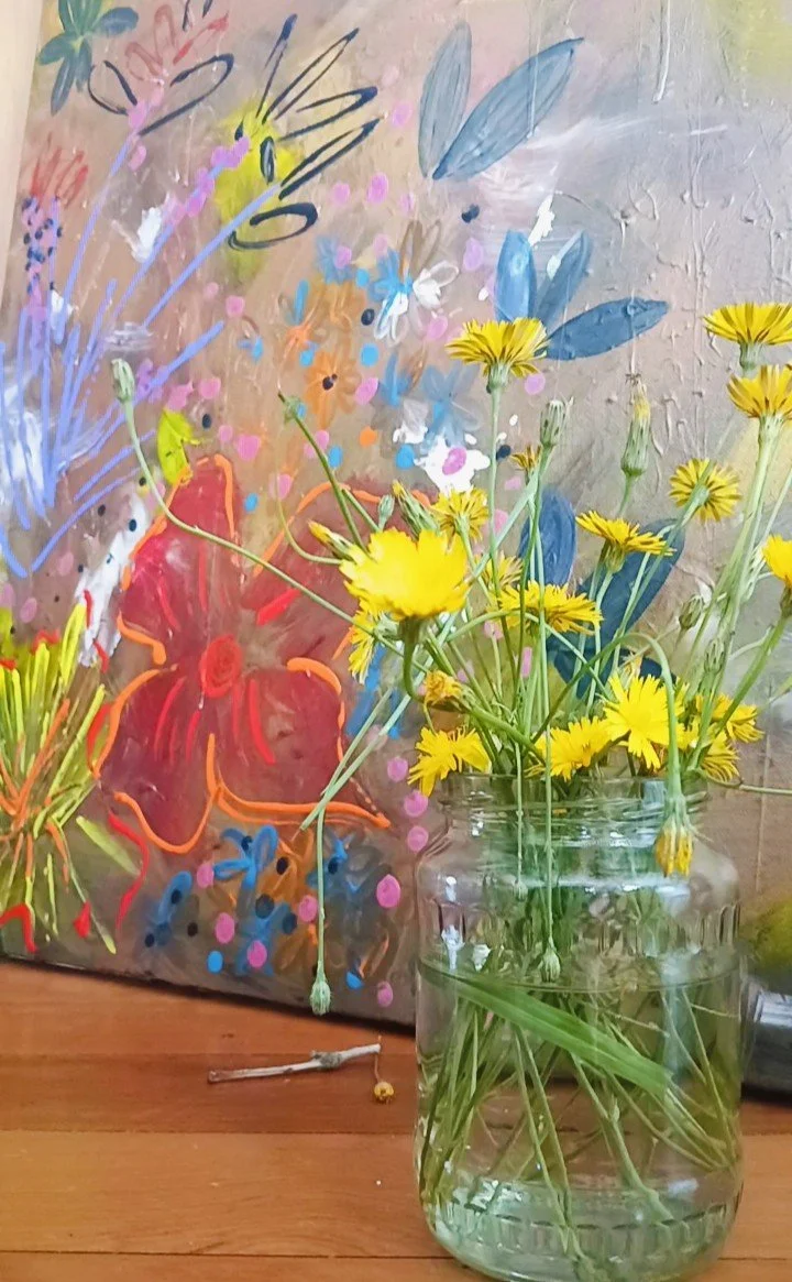 A glass jar filled with yellow daisies on a wooden surface, with a colorful, abstract floral painting in the background.