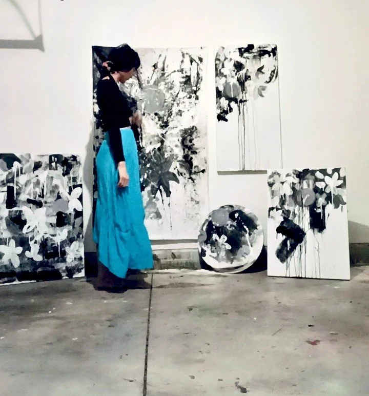 Woman standing in front of abstract black and white paintings displayed on a wall and on the floor, wearing a black top and a long bright blue skirt.