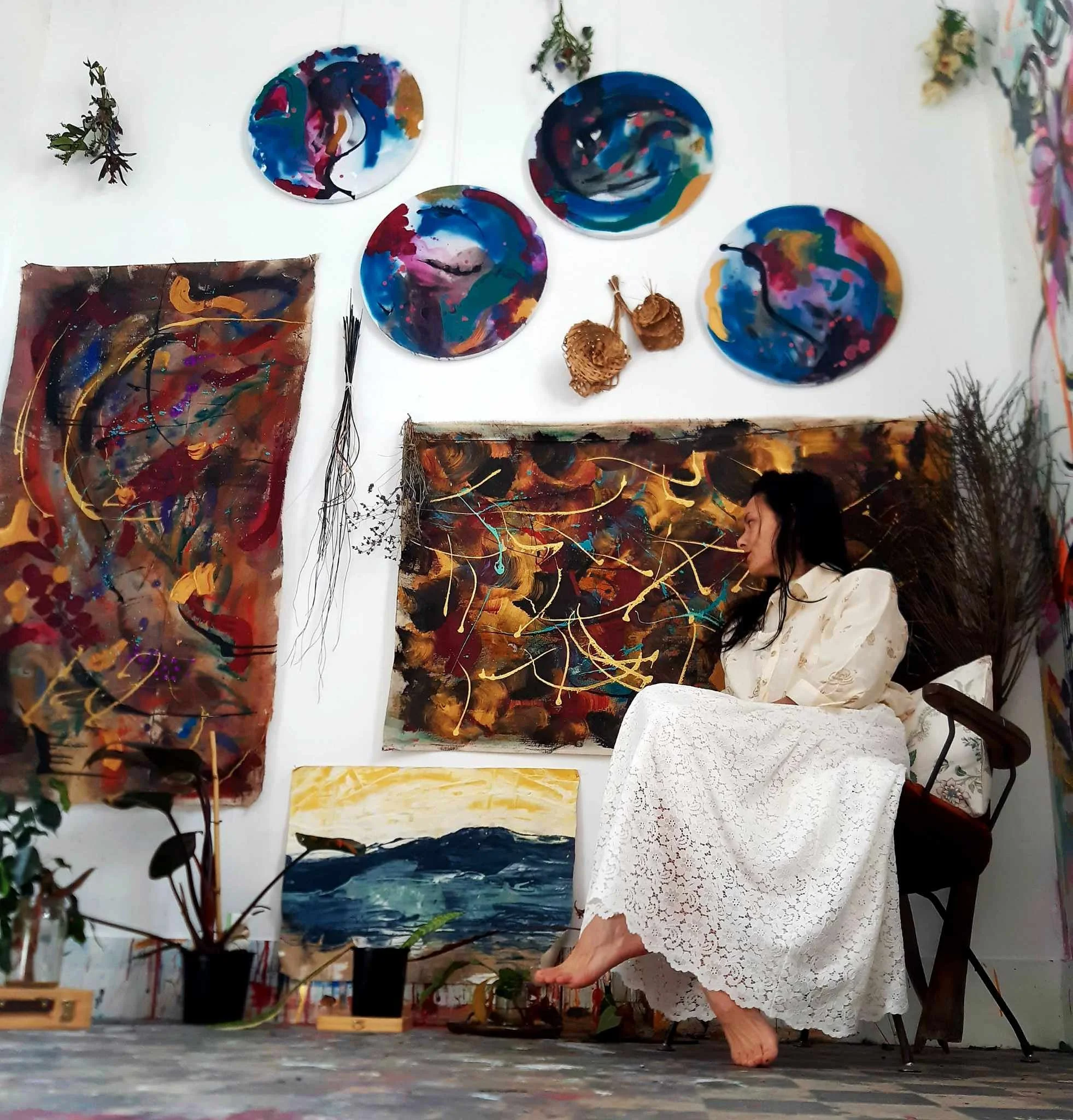 Abstract paintings on white walls, a woman in a white lace dress tears paper with her toes, plants and floral arrangements are placed around, colorful artwork is visible.
