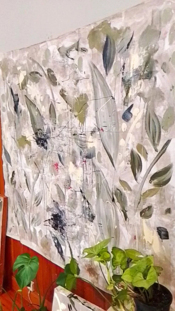 An abstract painting on a large canvas with neutral, green, and dark hues, leaning against a wood-paneled wall. In front of the painting, there are green leafy houseplants in pots.