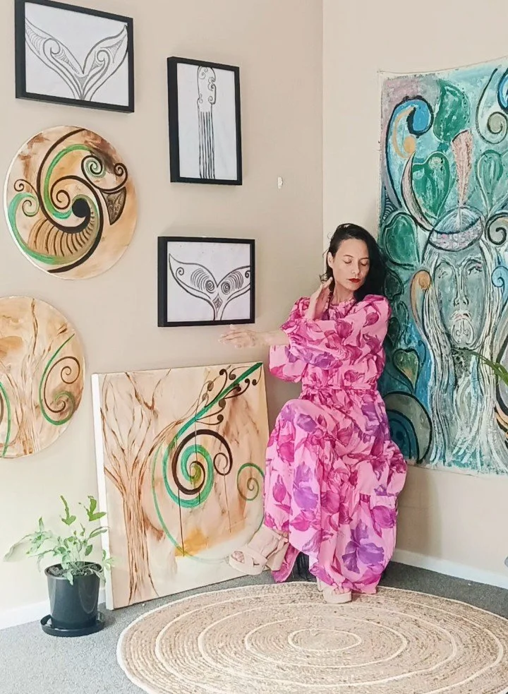 A woman in a pink floral dress is sitting against a beige wall, surrounded by abstract artistic paintings with swirling patterns, some framed and some on canvas. She is touching her neck with her eyes closed.