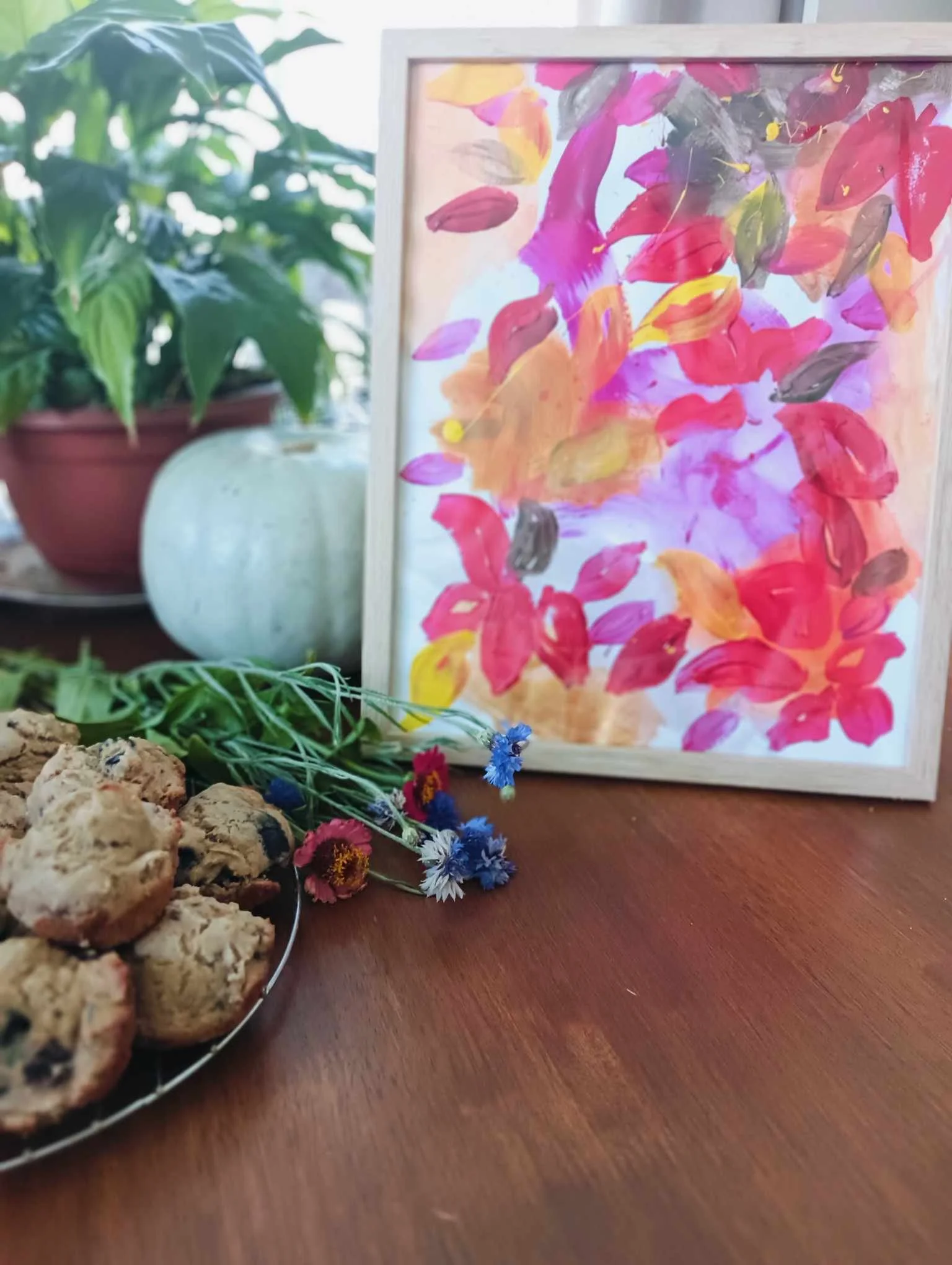 Photo of a table with homemade cookies, colorful dried flowers, a potted plant, a white decorative object, and a colorful abstract painting in a light wooden frame.