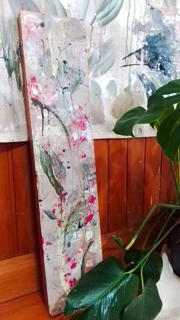 Colorful abstract floral painting on a tall rectangular canvas leaning against a wooden wall, with green leaves from a houseplant partially in front.