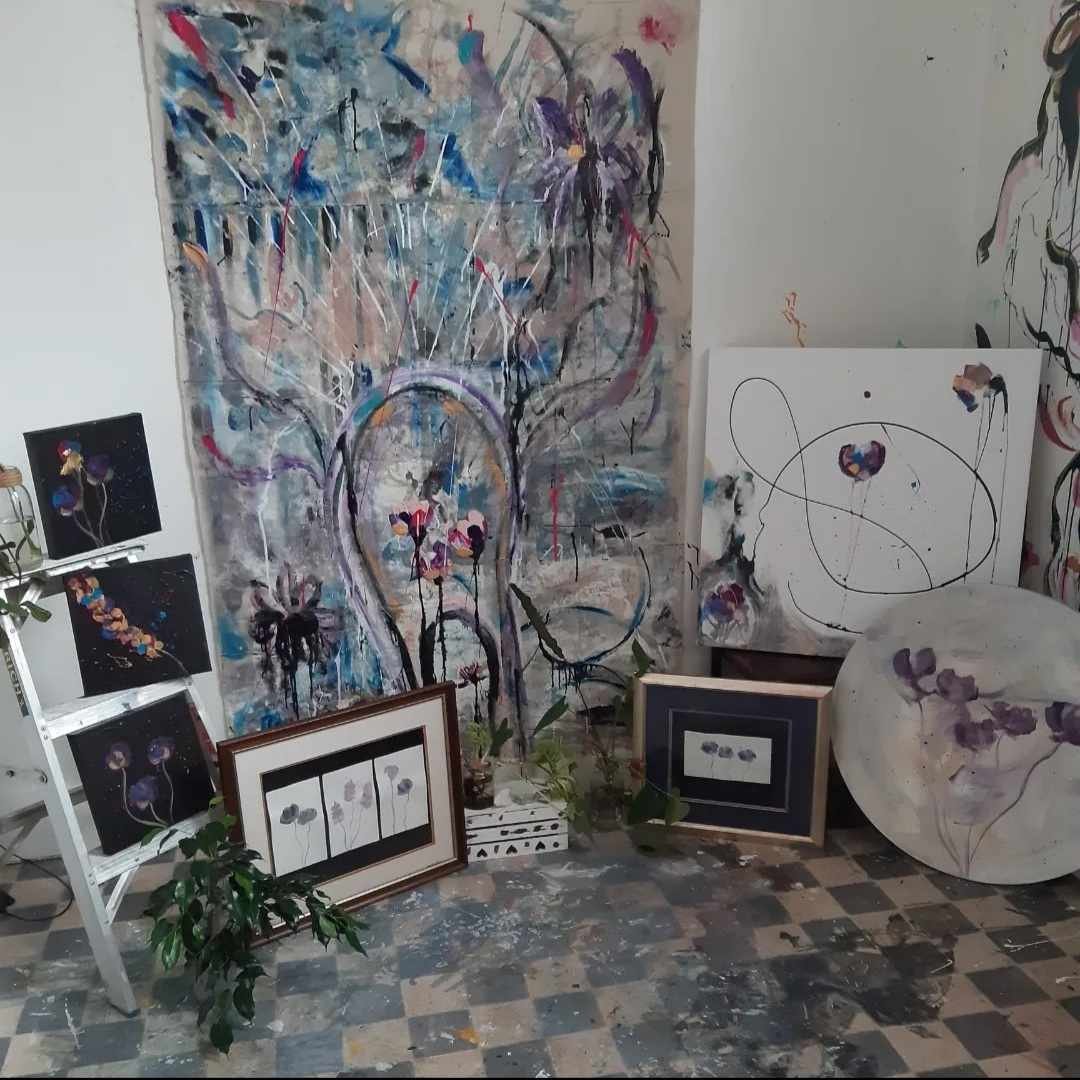 An art studio with several paintings, including a large abstract floral artwork on the wall, smaller framed floral paintings on the floor, and works on a white ladder and circular canvas, with paint splatters on the floor.