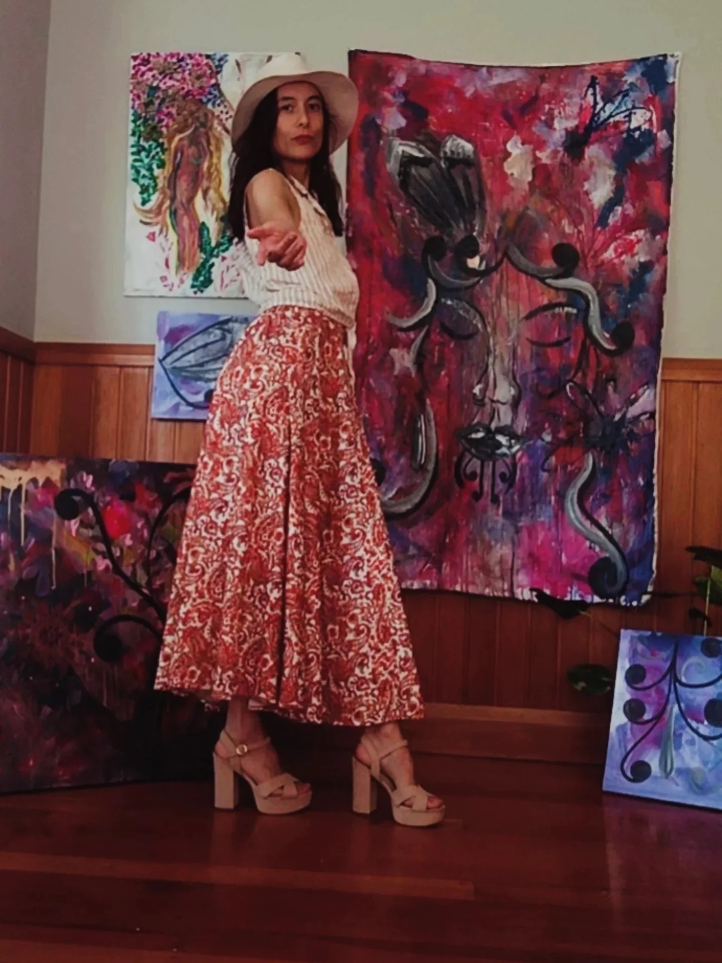 A woman standing in front of colorful abstract paintings, wearing a wide-brimmed hat, sleeveless striped top, long patterned skirt, and beige platform shoes. She is pointing with her right hand.