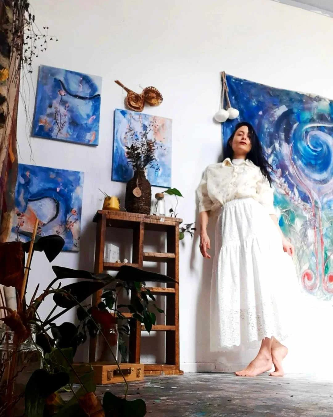 A woman in a white dress standing barefoot in an artist's studio, with abstract blue paintings on the walls and various plants and decor, including a large swirling artwork behind her.