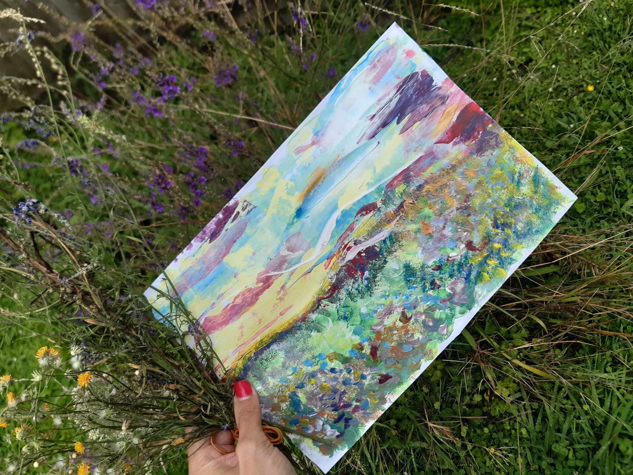 A person's hand holding a colorful abstract painting with brushstrokes of purple, blue, yellow, and green, against a background of grassy plants and purple flowers.