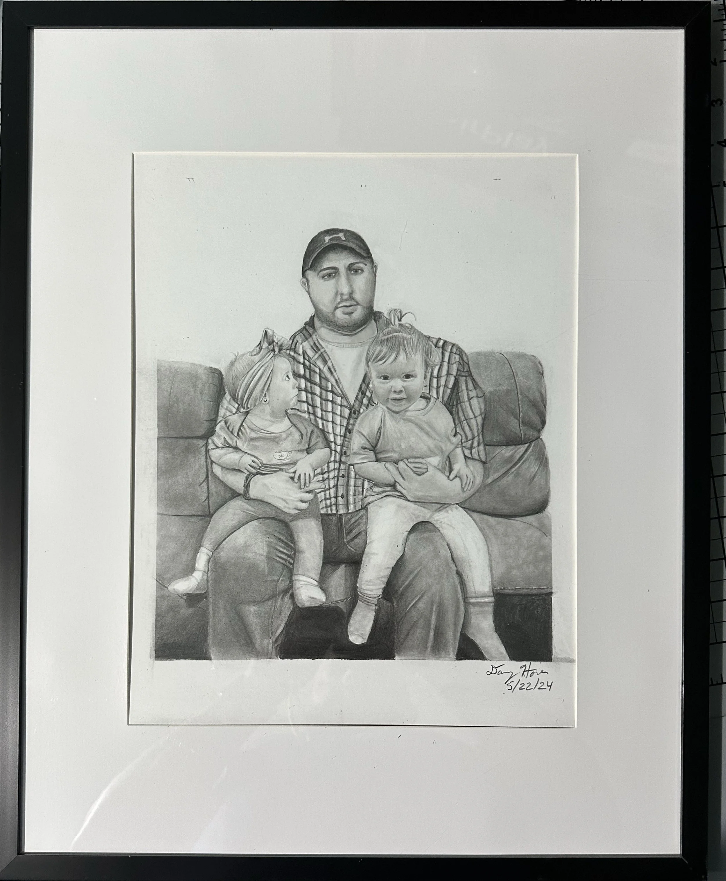 A black and white drawing of a man sitting on a couch holding two children. One child is on his lap and the other is sitting beside him. The man is wearing a plaid shirt and a cap. Coats are visible on the couch's armrest. The drawing is signed and d