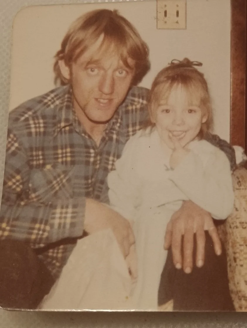 A man with blond hair and a young girl with blond hair, smiling, sitting together indoors. The man is wearing a plaid shirt and the girl is wearing a light-colored sweater. They are sitting close to each other, with the man's hand resting on the girl