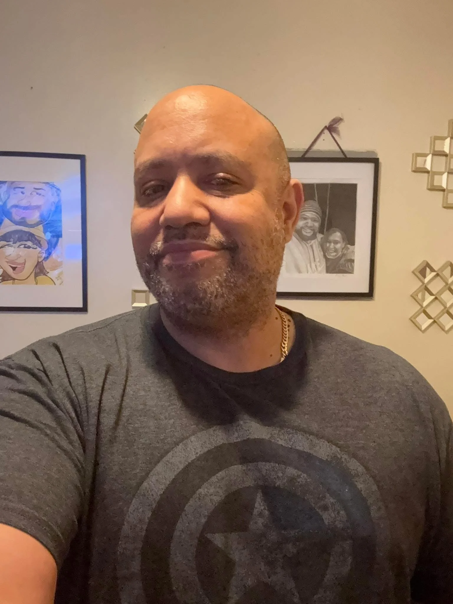 A man with a bald head and facial hair taking a selfie in front of wall art and framed pictures.