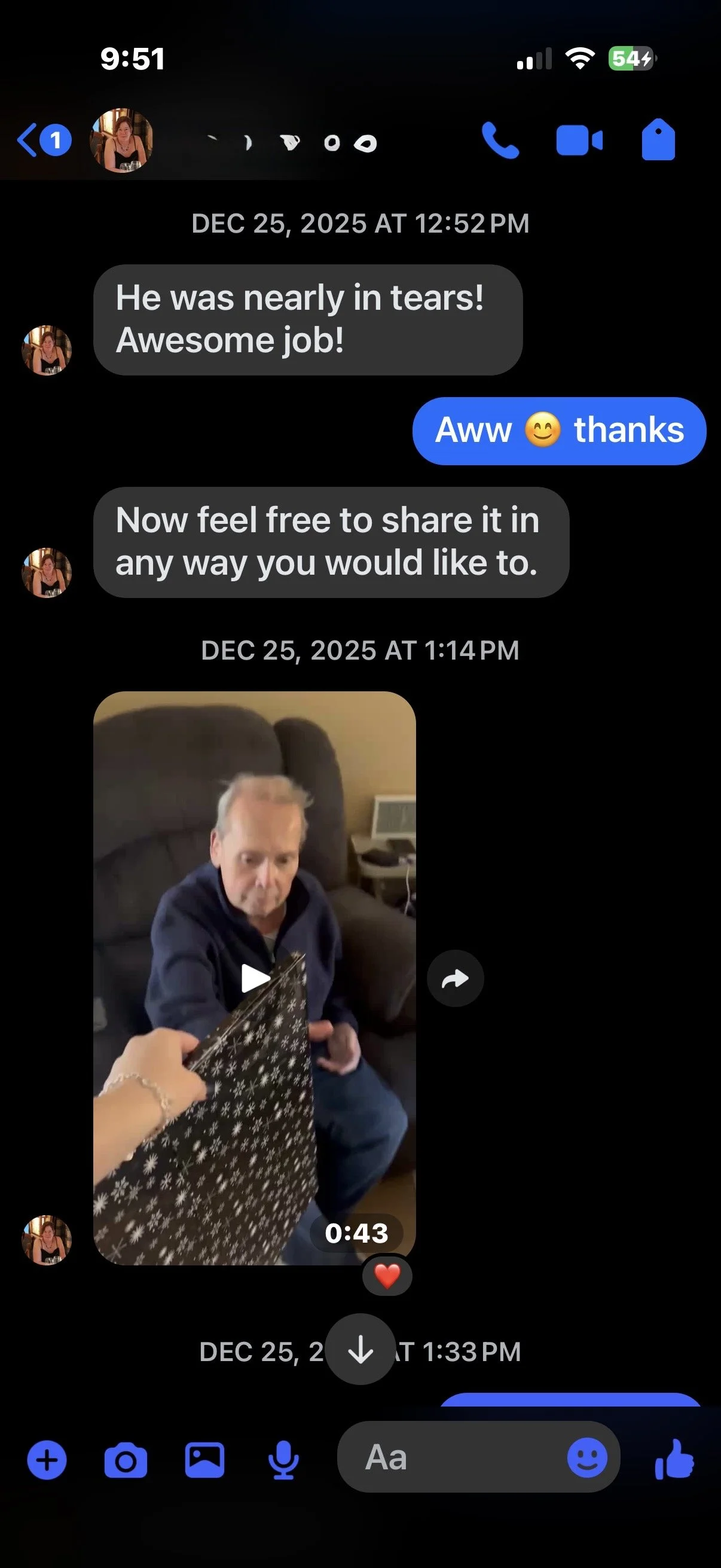 A video chat screenshot showing an older man with gray hair sitting on a brown armchair, holding a Christmas gift bag with a snowflake pattern, while a woman presents it to him. The background includes a beige wall and a small table with an electronic device.