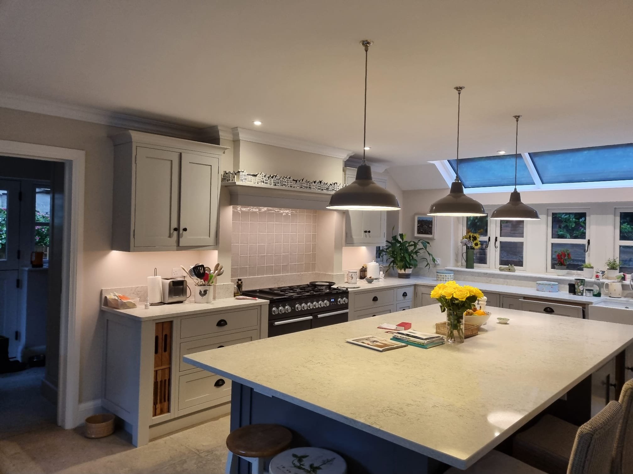 A spacious kitchen with a large island, white cabinets, pendant lighting, and a skylight, decorated with plants and flowers.