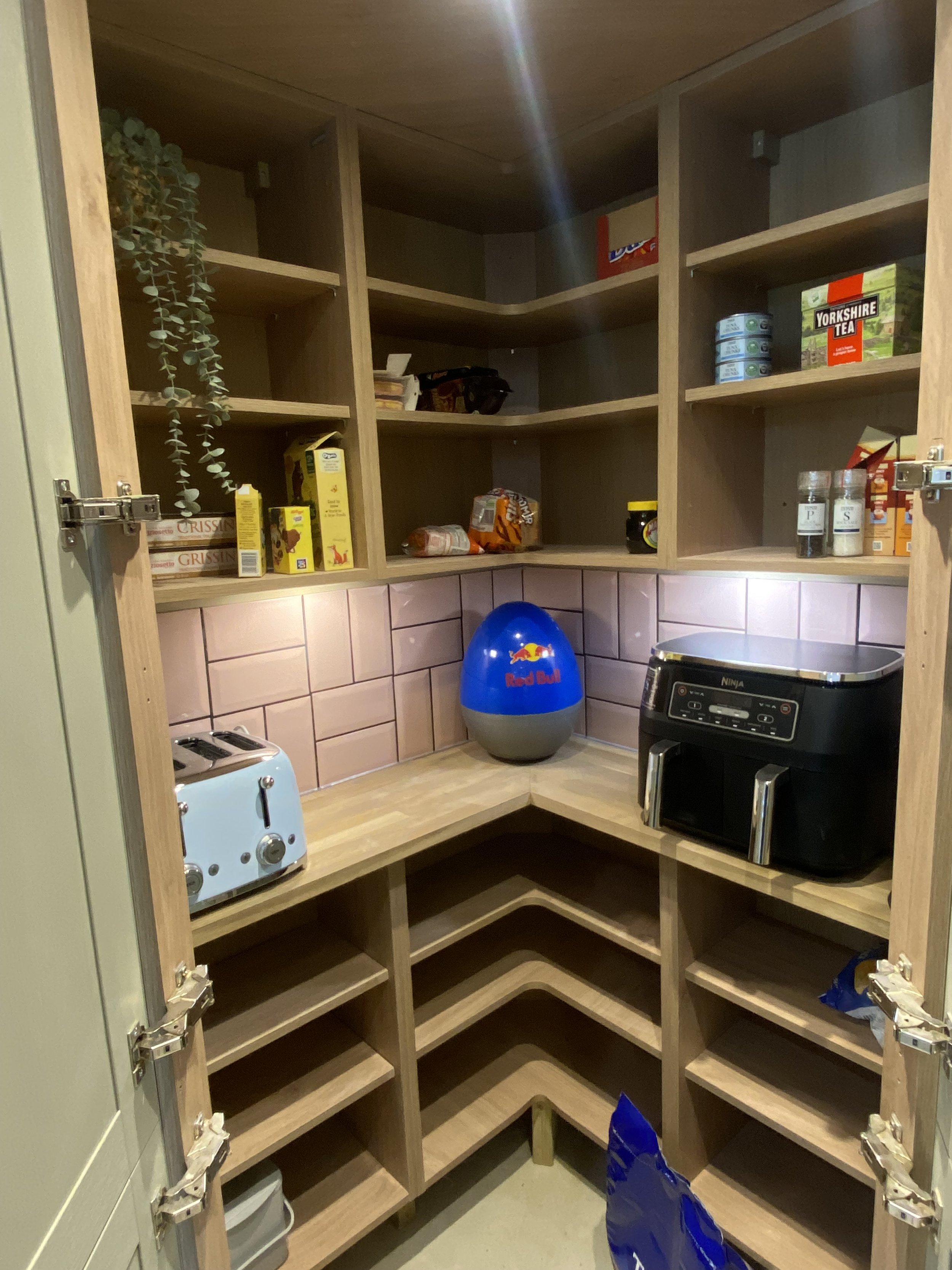 An empty kitchen pantry with wooden shelves and some stored items including cereal boxes, canned goods, a Red Bull themed mini fridge, a toaster, and a Ninja appliance.