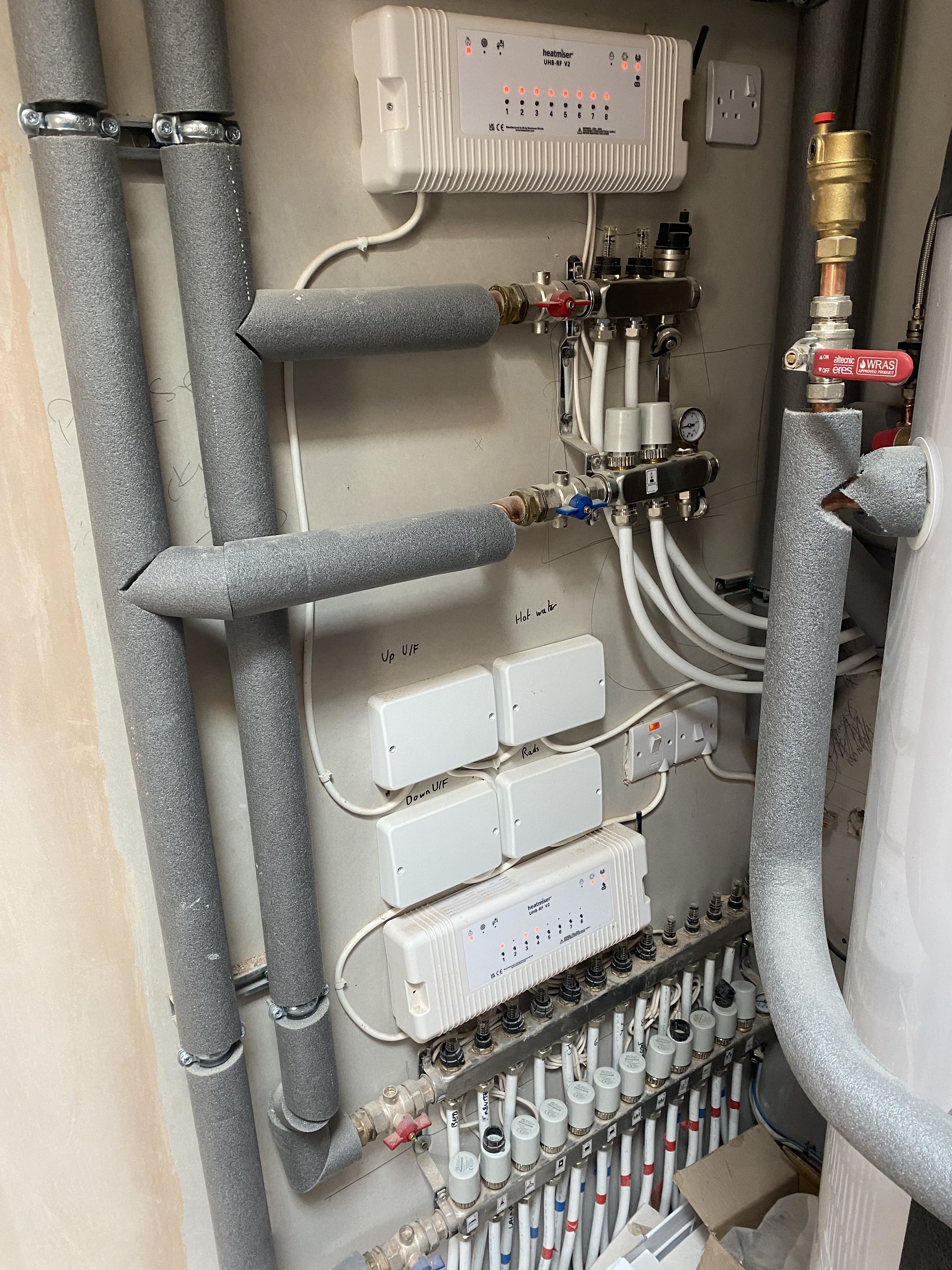 A plumbing and heating system with pipes, valves, gauges, and electronic controllers mounted on a wall, including insulated pipes, a heat management unit, and various control switches.