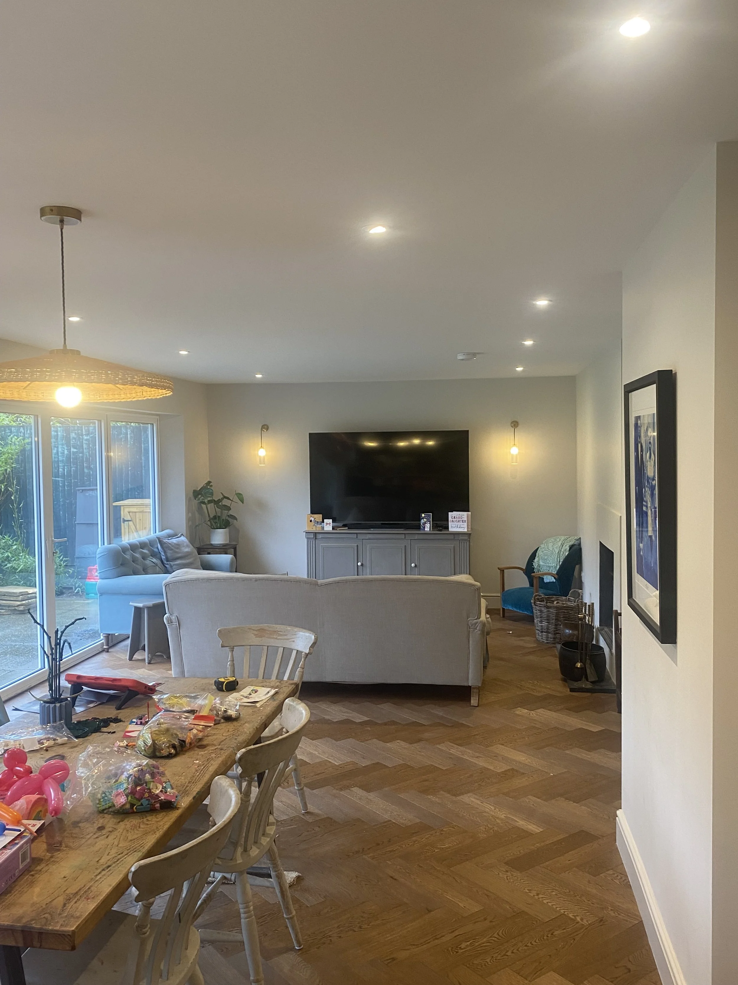 A cozy living room with a large flat-screen TV on a gray cabinet, a beige patterned sofa, a white armchair, and a sliding glass door leading outside. The room has wooden flooring, wall-mounted lights, and a dining table scattered with toys and craft 