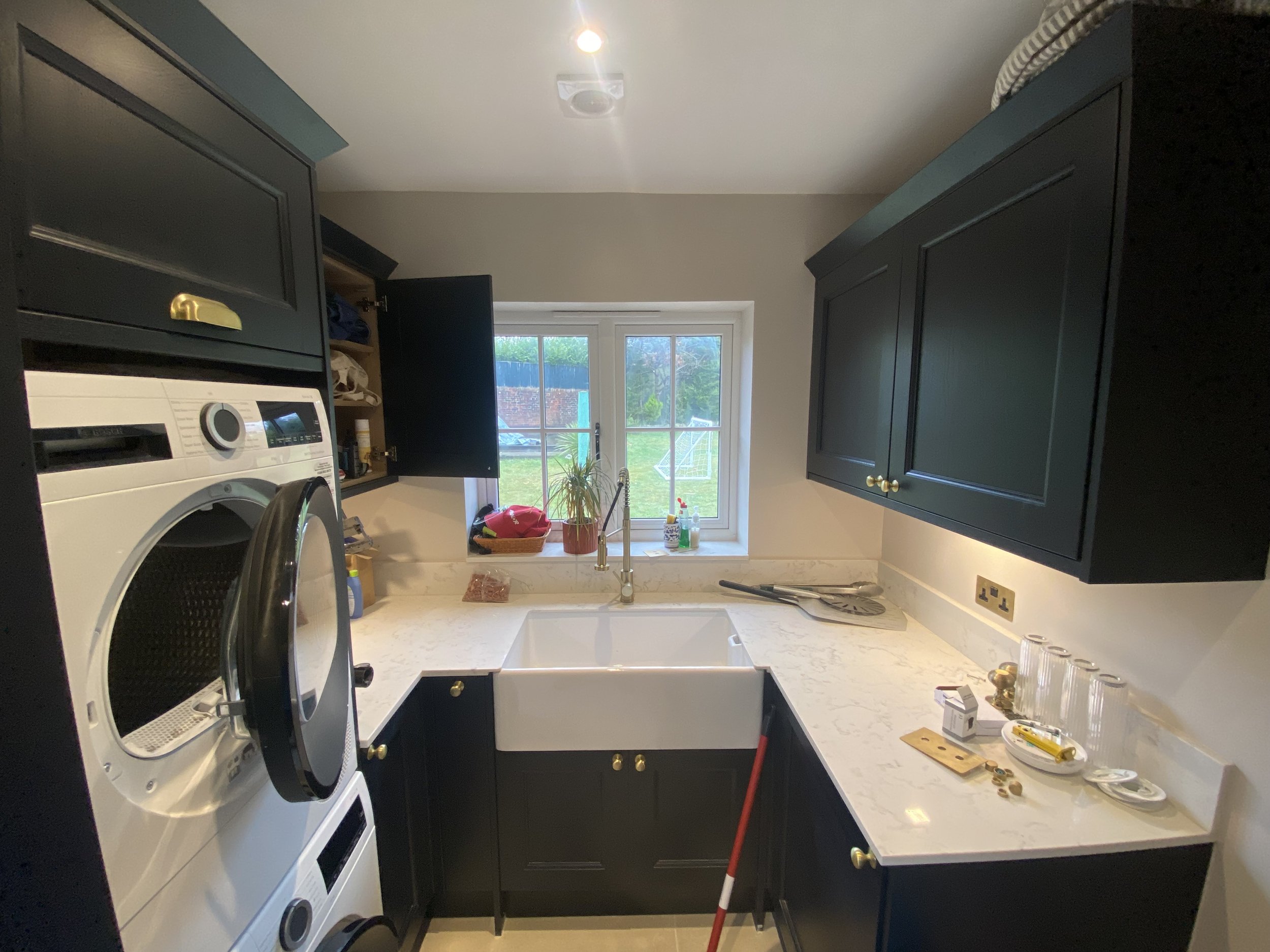 A laundry room with dark cabinets, a washing machine, a sink, a window, and various laundry supplies.