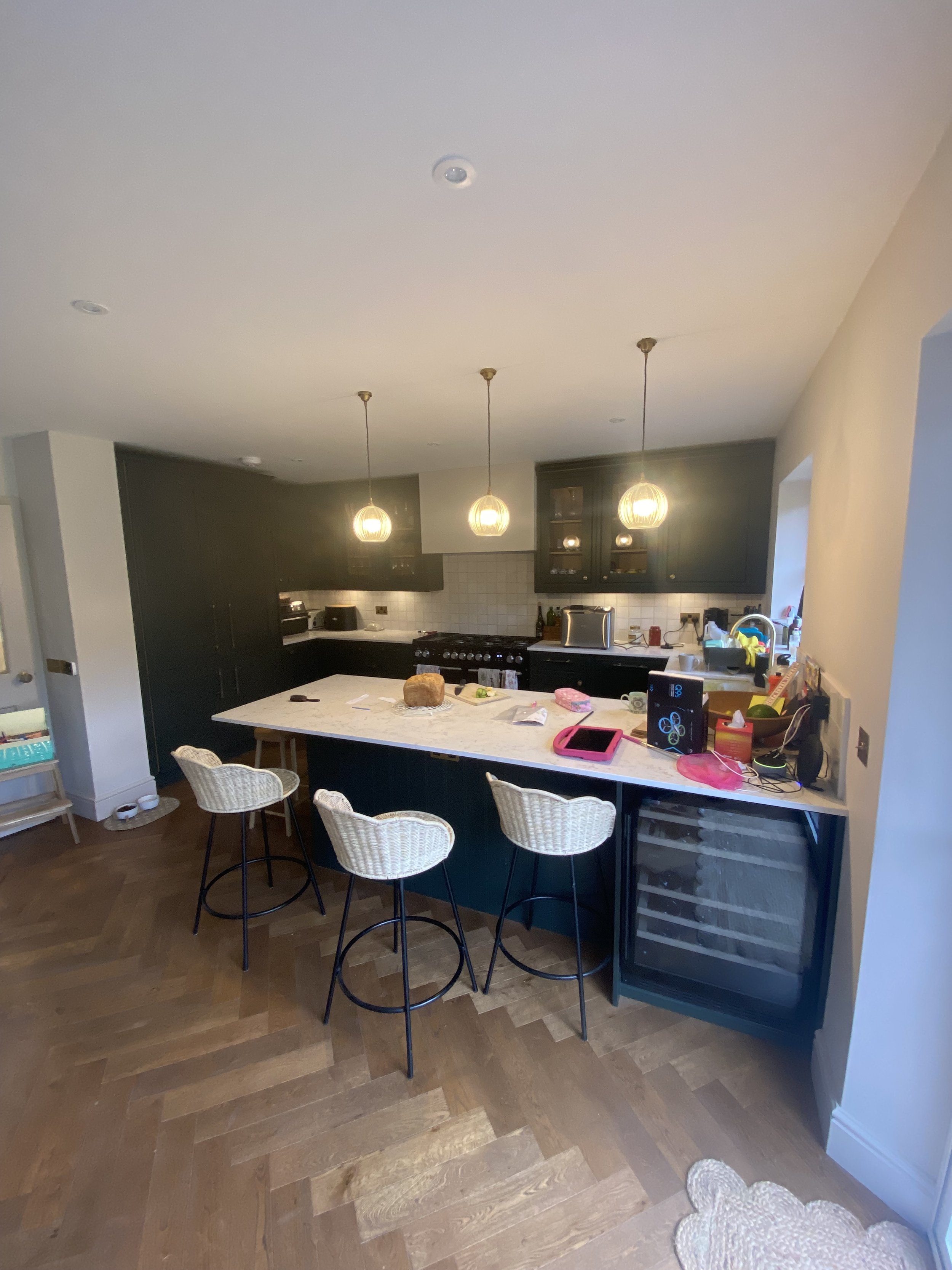 A kitchen with black cabinets, a large white marble island, three hanging pendant lights, a stovetop, a wine cooler, and various items on the counter including a loaf of bread and a pink tablet. There are three white wicker chairs around the island, 